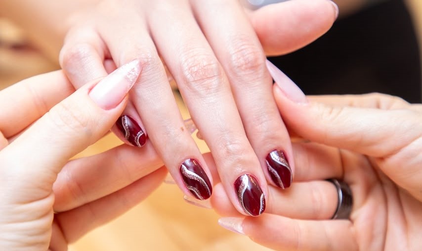 Mãos de uma cliente com esmaltação em gel vinho e detalhes em glitter, sendo preparadas por uma profissional